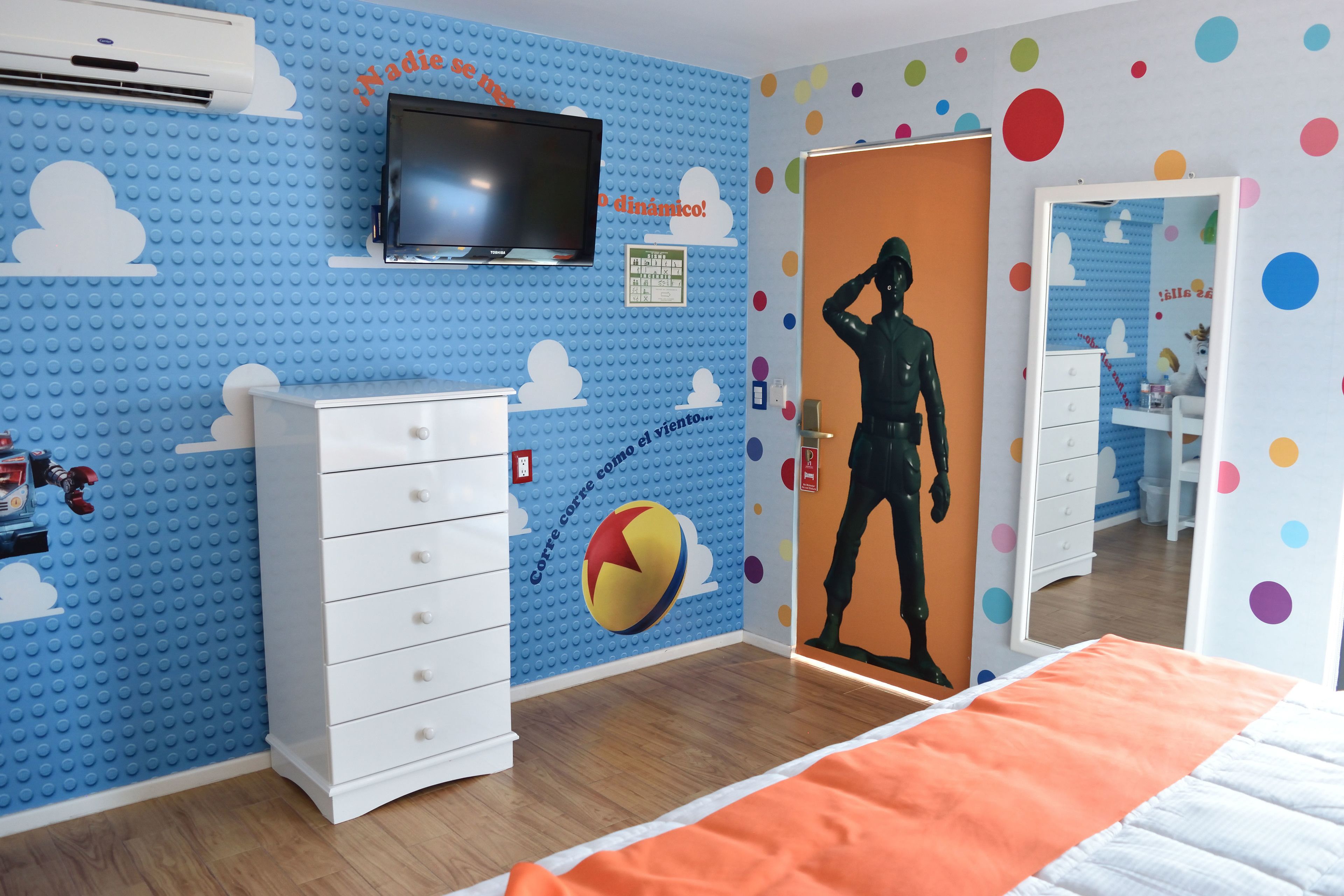 Una habitación con una pared azul de LEGO, un tocador blanco, una cama con una manta naranja y una puerta con un estampado colorido de lunares.