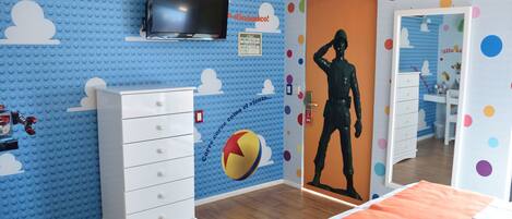 Una habitación con una pared azul de LEGO, un tocador blanco, una cama con una manta naranja y una puerta con un estampado colorido de lunares.