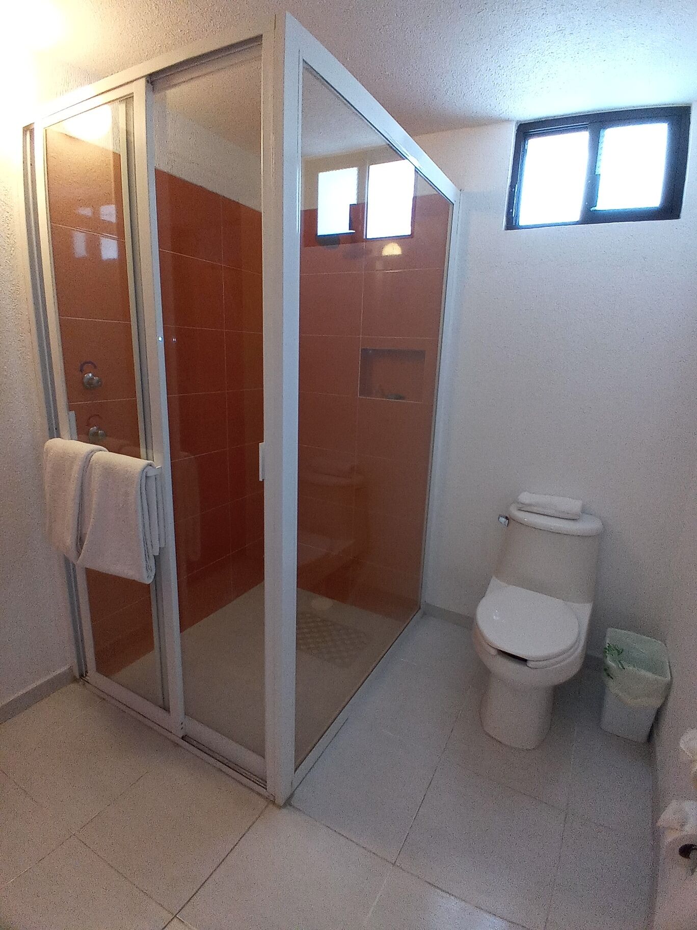 Un baño con mampara de vidrio, inodoro y una ventana pequeña.