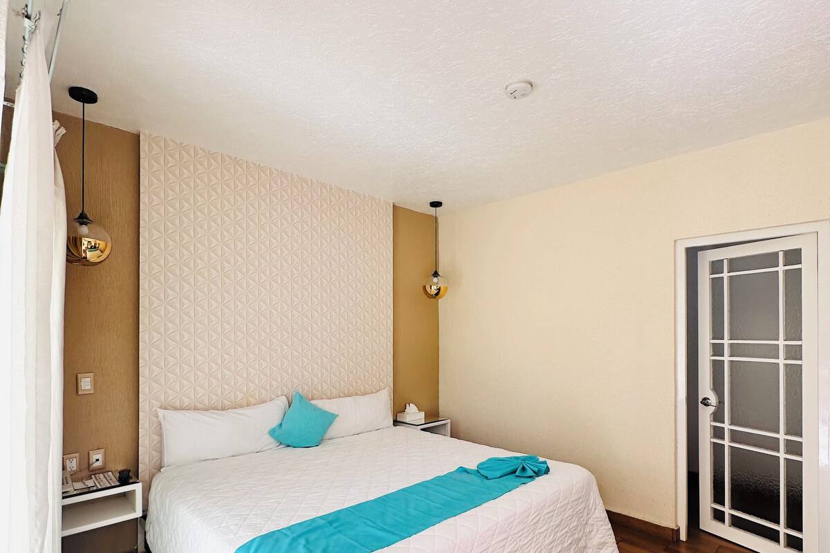 Habitación de hotel ordenada con una cama grande, piso de madera, cabecera blanca y una puerta con paneles de vidrio esmerilado.