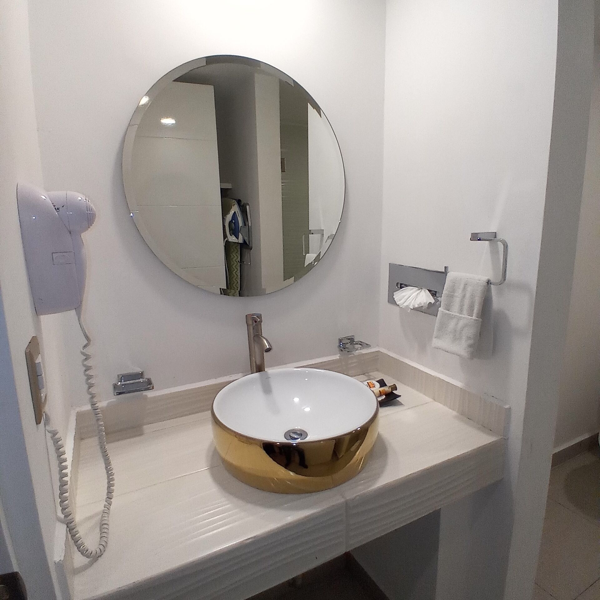 Une salle de bain avec un miroir rond, un lavabo, un sèche-cheveux et un porte-serviettes.