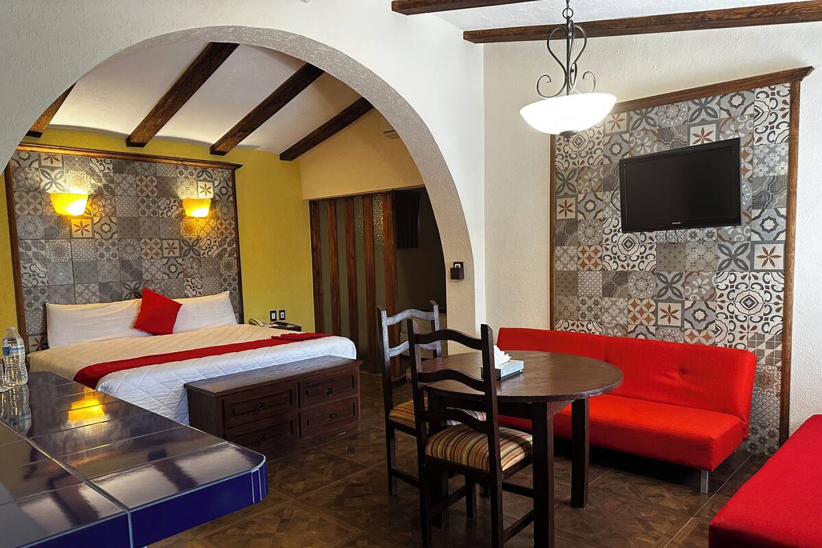Una habitación de hotel con cama, una pequeña mesa de comedor, un sofá rojo y un televisor instalado en la pared.