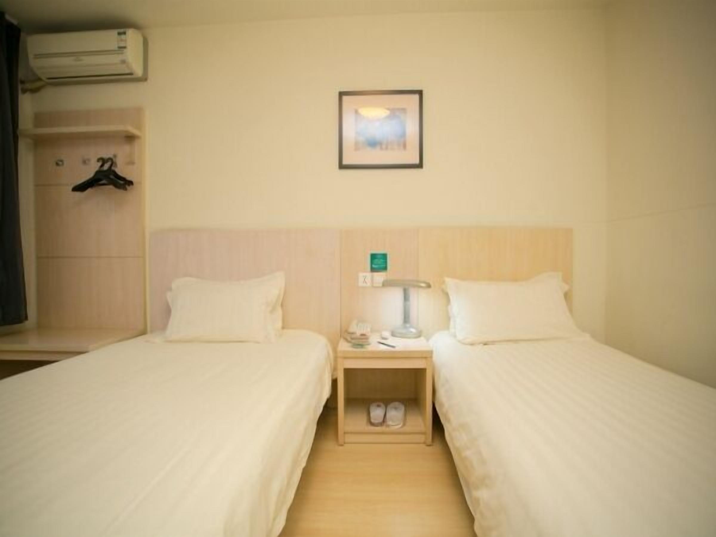 A hotel room with two single beds, a nightstand with a lamp, a small shelf with shoes, and a framed picture on the wall.