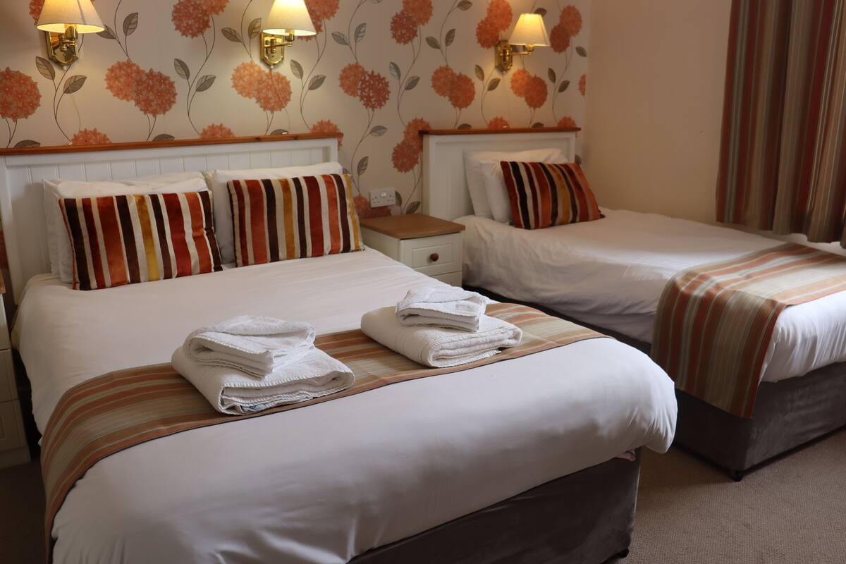 Ein Hotelzimmer mit zwei Betten, jeweils mit gestreifter Bettwäsche und Kissen, jeweils einem hölzernen Kopfteil, einem Nachttisch und einer Wand mit floralem Wandpapier.