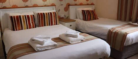 Ein Hotelzimmer mit zwei Betten, jeweils mit gestreifter Bettwäsche und Kissen, jeweils einem hölzernen Kopfteil, einem Nachttisch und einer Wand mit floralem Wandpapier.