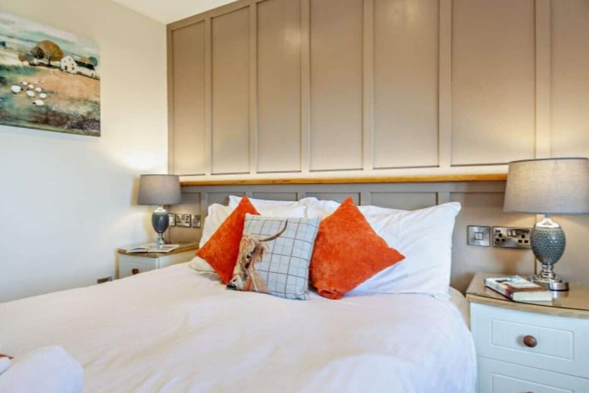 Ein ordentlich bezogenes Bett mit weißen Leinentüchern und zwei orangefarbenen Kissen, ein Nachttisch mit Lampe sowie ein Bild an der Wand.