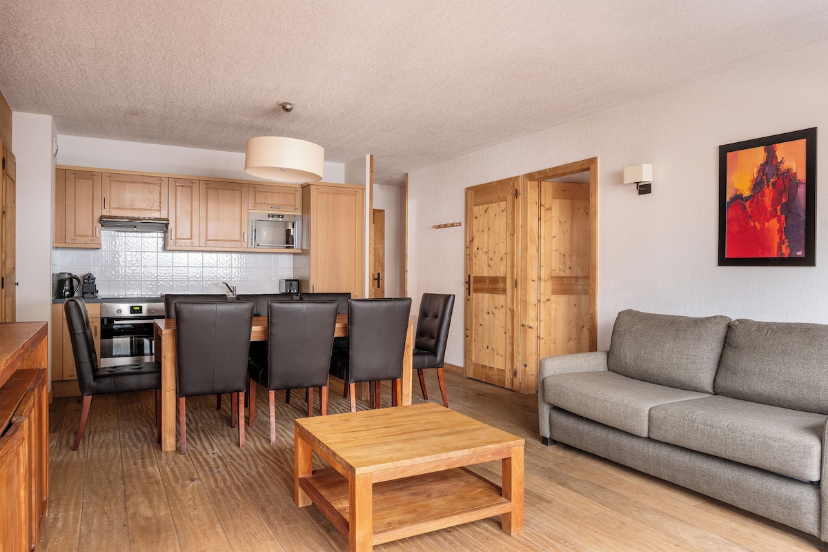 A modern kitchen with wooden cabinets, a dining table with chairs, a grey sofa, and a painting on the wall.
