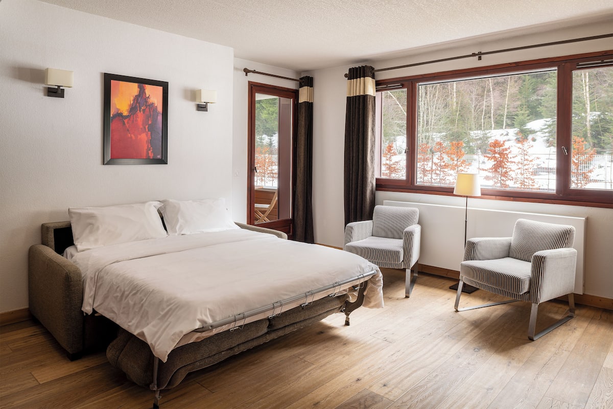 A bedroom with a bed, a chair, a window with curtains, and a painting on the wall.