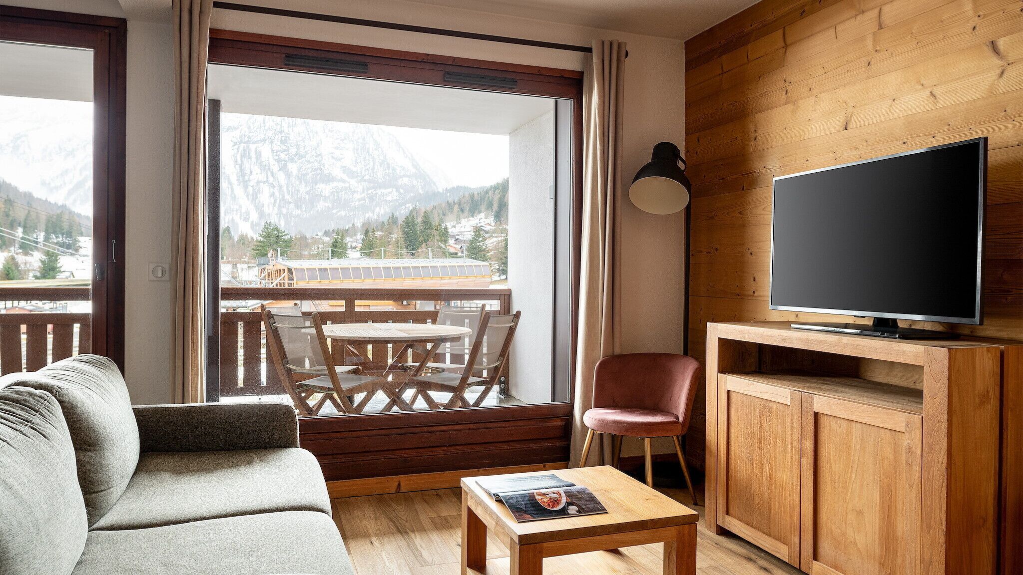 Una sala de estar acogedora con un sofá, una mesa de centro, un televisor sobre un mueble de madera, y un balcón con una mesa y sillas.