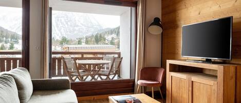Una sala de estar acogedora con un sofá, una mesa de centro, un televisor sobre un mueble de madera, y un balcón con una mesa y sillas.