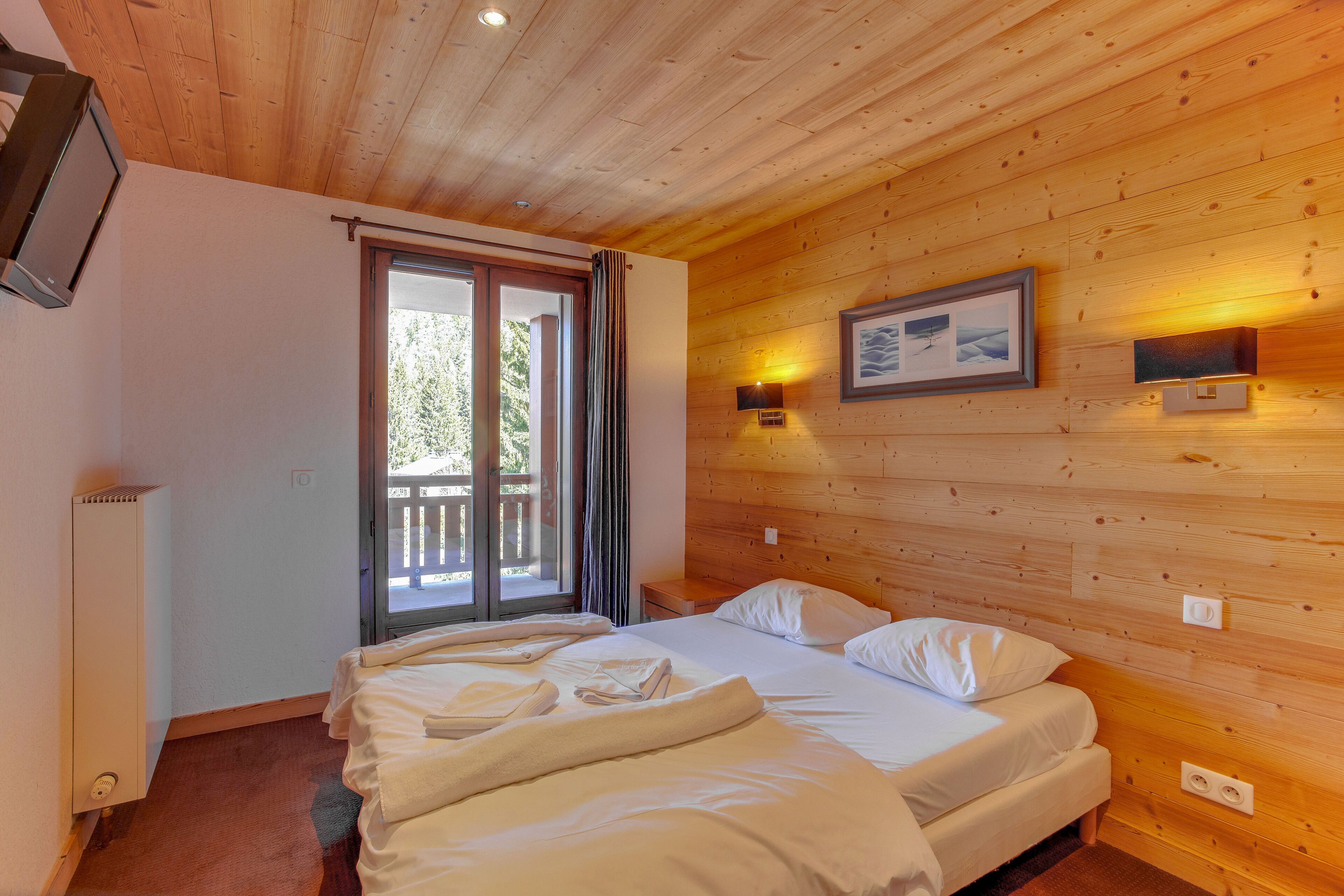 A bedroom with a wooden ceiling, a bed with white linens, a window with curtains, and a wall-mounted light fixture.