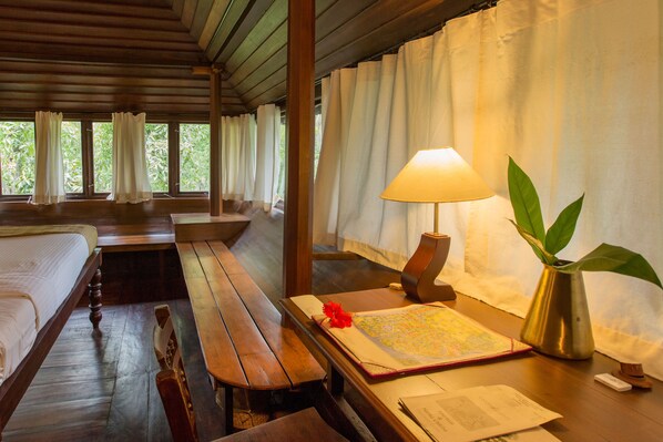 Una habitación con una cama de madera, un banco de madera, una mesa con un mapa y papeles, una lámpara y una planta en maceta.