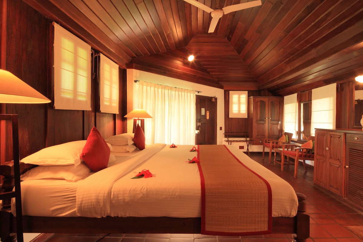 A hotel room with a large bed, wooden ceiling, and a dining area with chairs and a table.