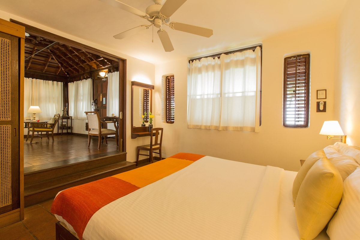 A hotel room with a bed, a ceiling fan, a window with curtains, a desk with a chair, and a mirror.
