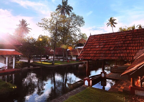 A traditional building with a red tiled roof, a small water body, and palm trees.