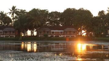 A row of bungalow-style buildings with a reflective water body in front, surrounded by palm trees.