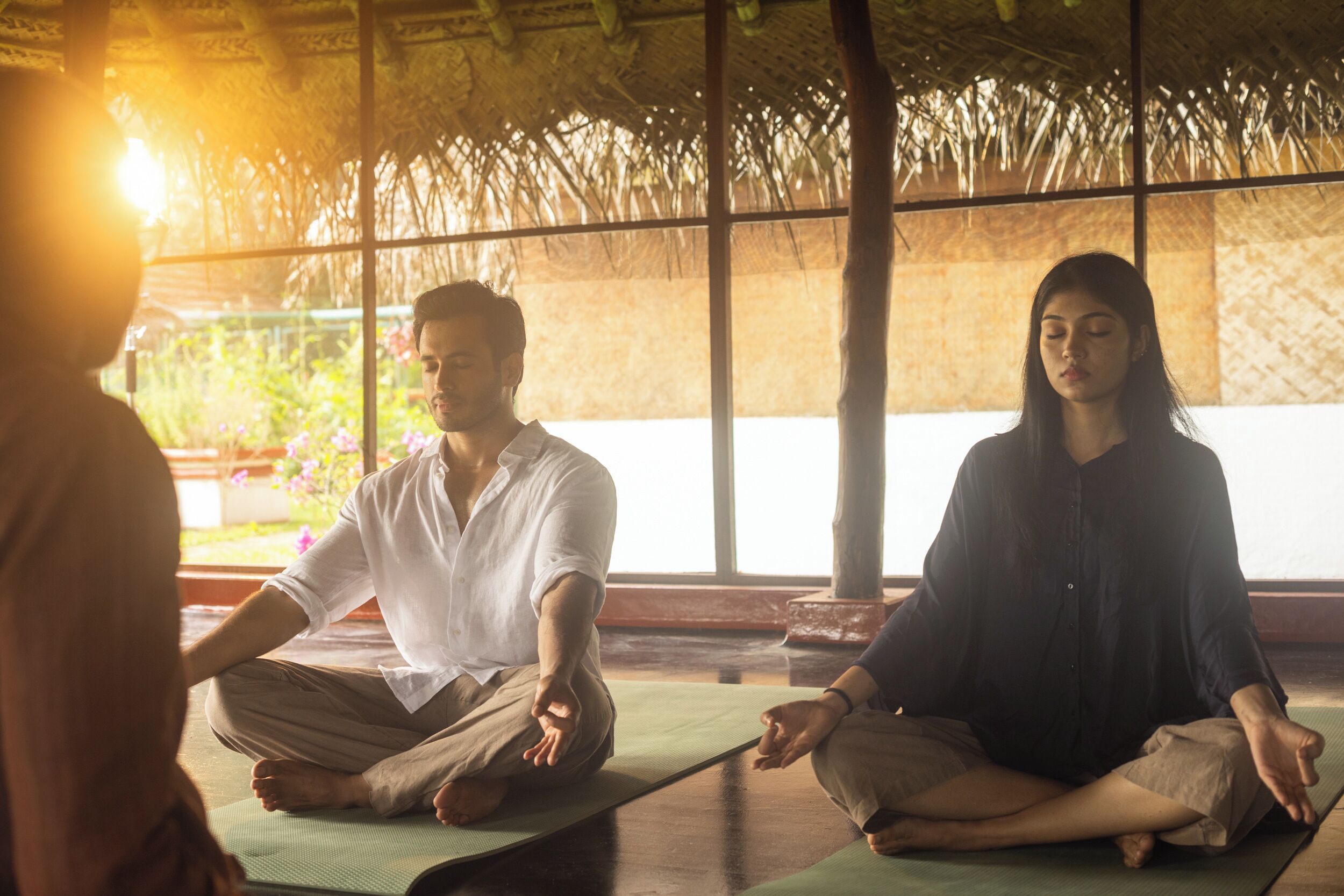 A group of people meditating in a room with a thatched roof and large windows.