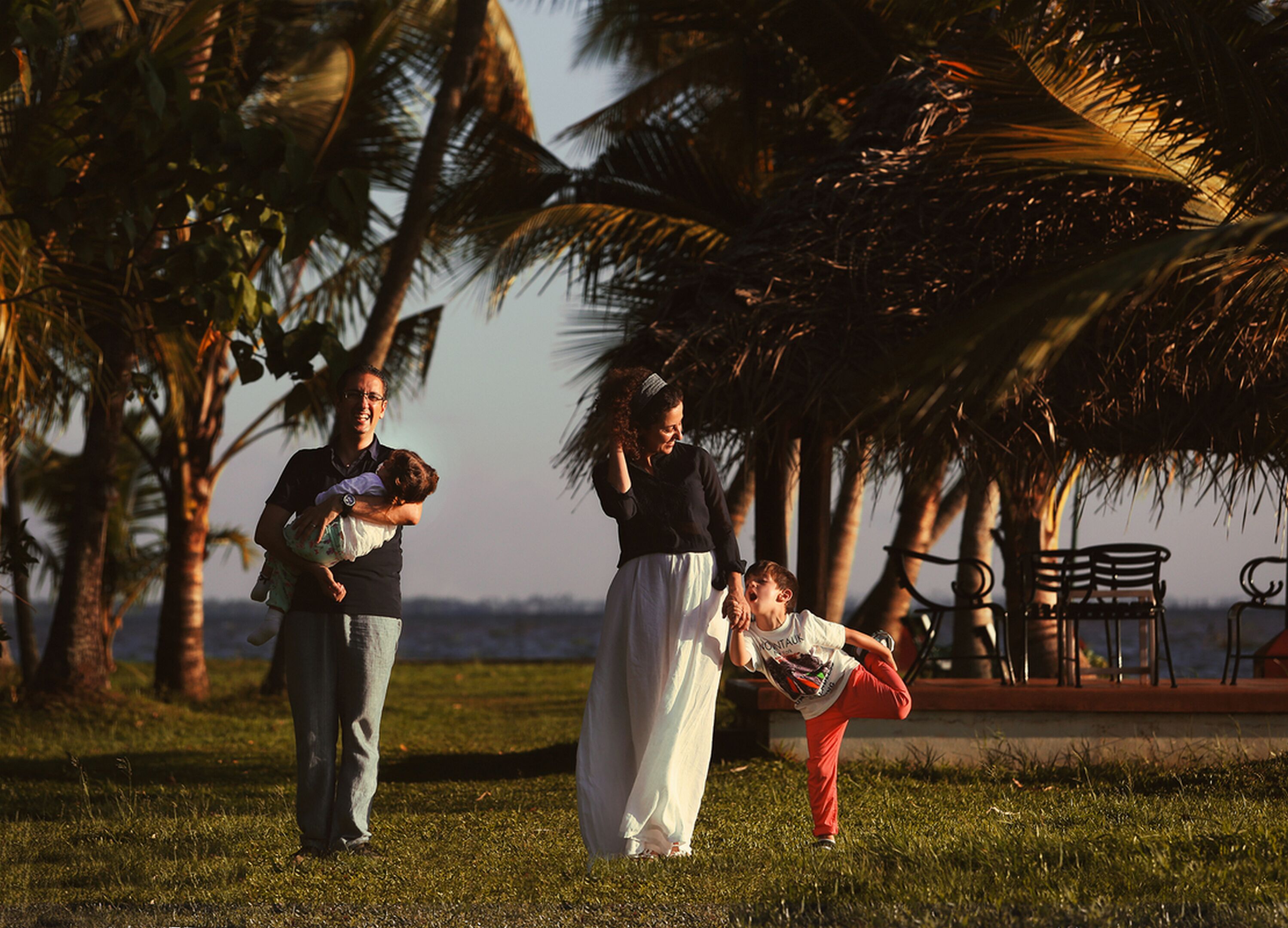 A family of three in a park with palm trees and a bench in the background.