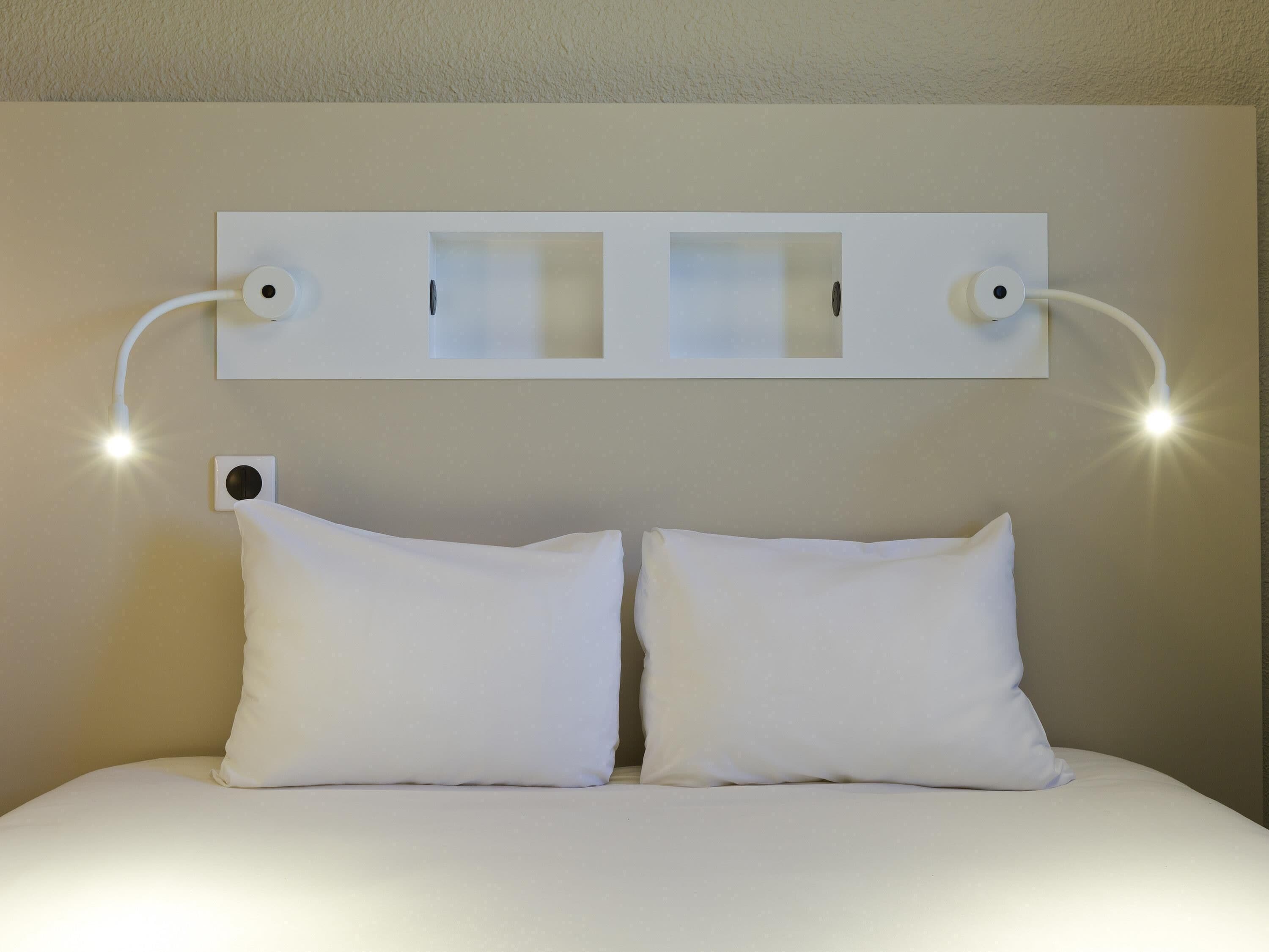 Habitación de hotel con cama, dos almohadas blancas, cabecera con iluminación integrada y lámpara de pared.