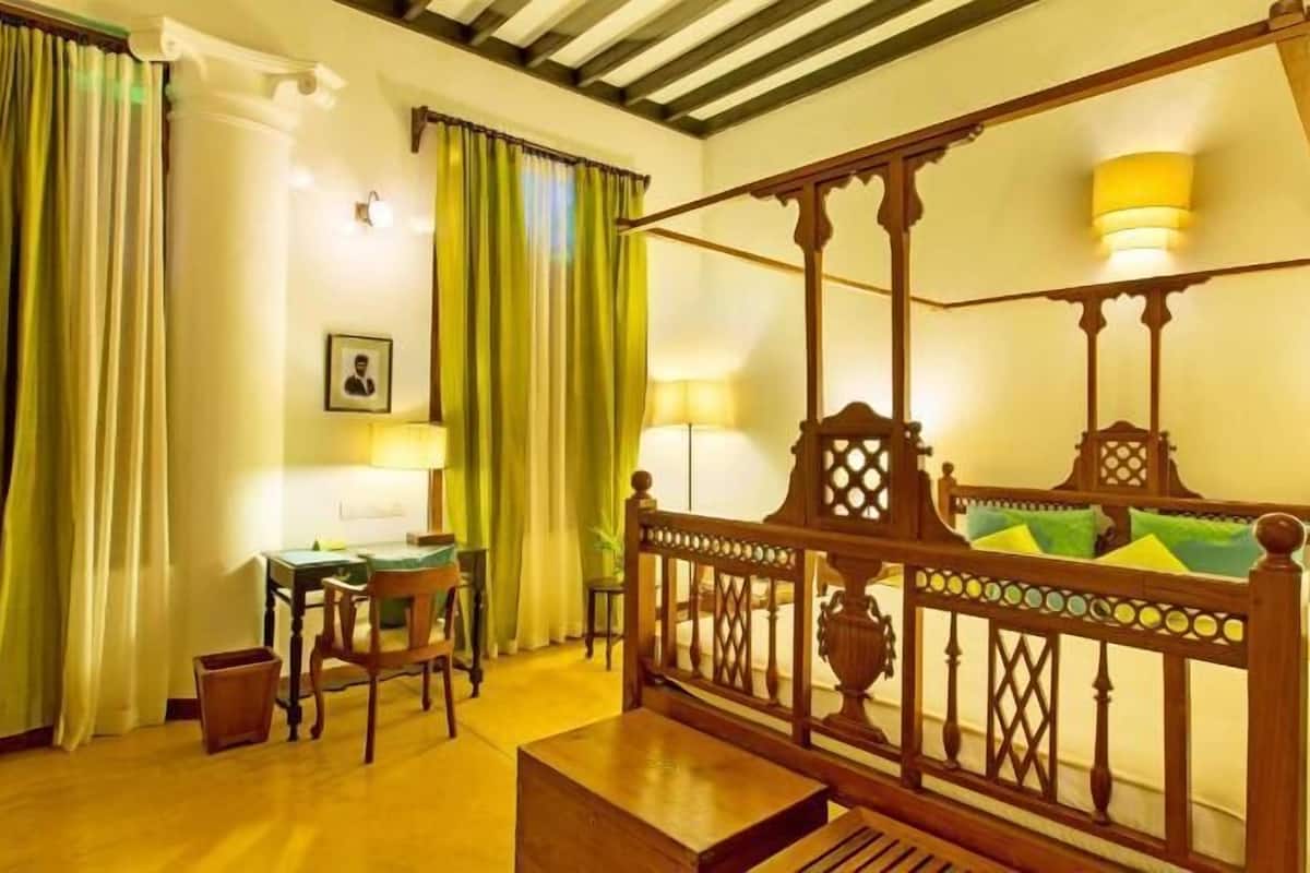 A room with a four-poster bed, a desk with a chair, a bedside table, and a lamp.