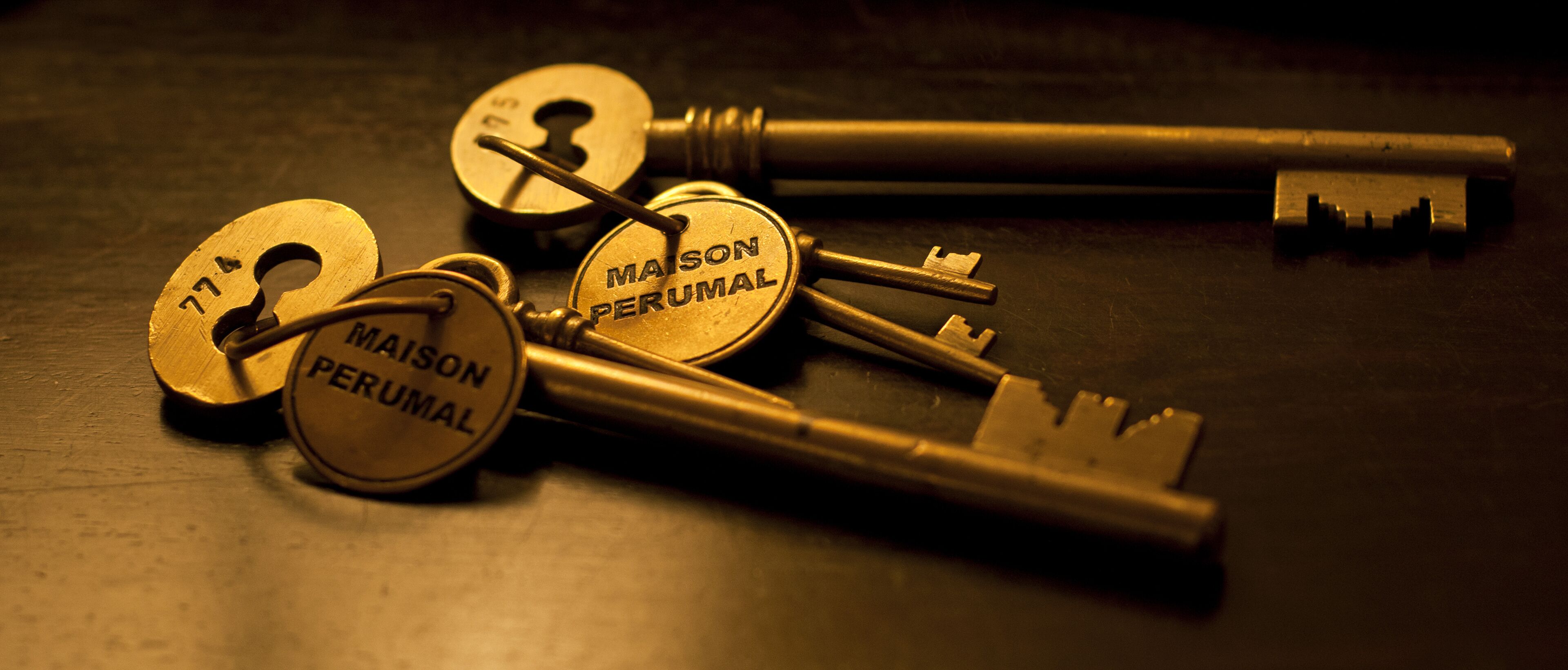 A set of keys with the inscription "MAISON PERUMAL" on them, lying on a dark surface.
