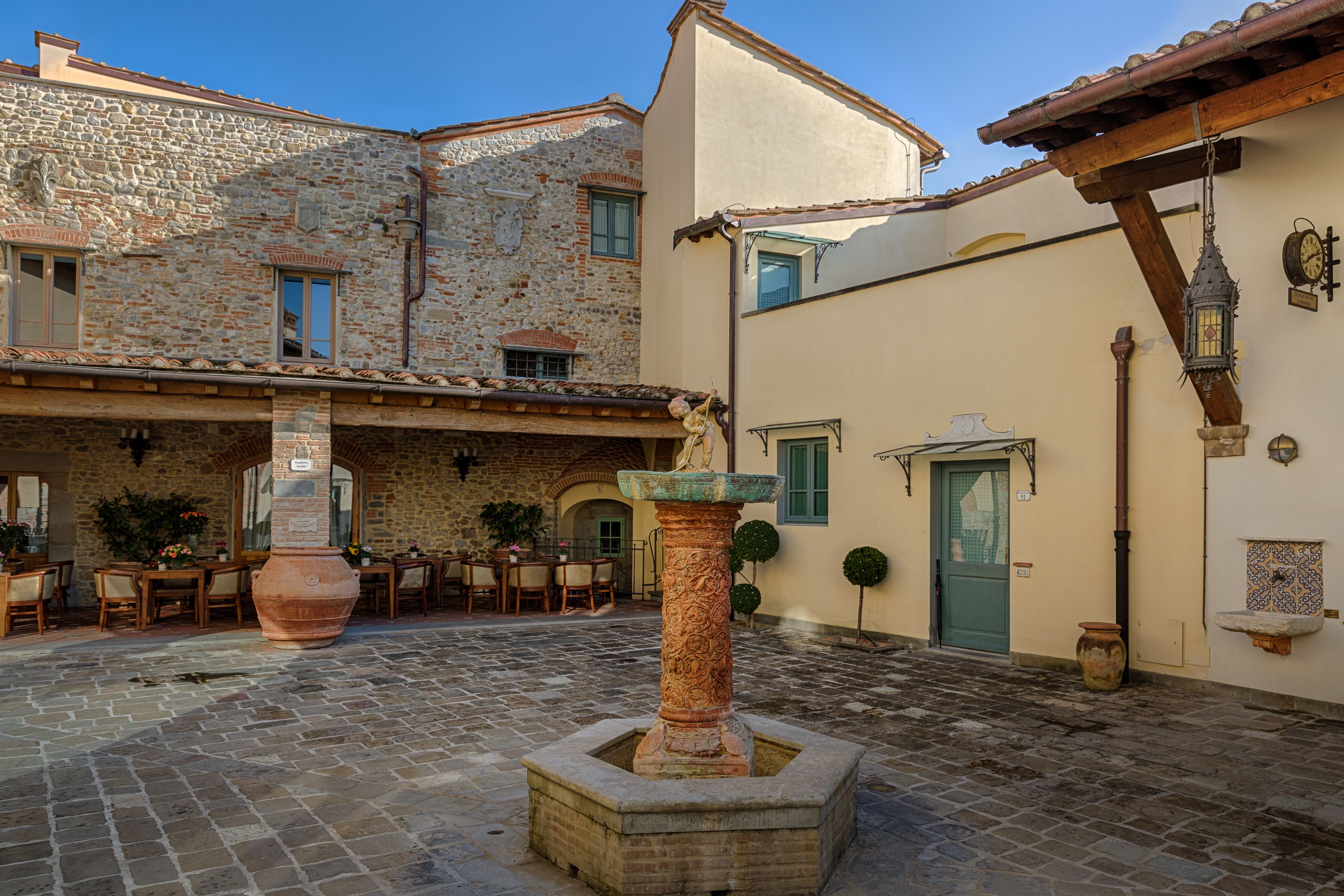 Un patio con suelo de piedra, una fuente en el centro, asientos exteriores y un edificio con una puerta y ventanas verdes.