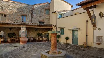 Un patio con suelo de piedra, una fuente en el centro, asientos exteriores y un edificio con una puerta y ventanas verdes.