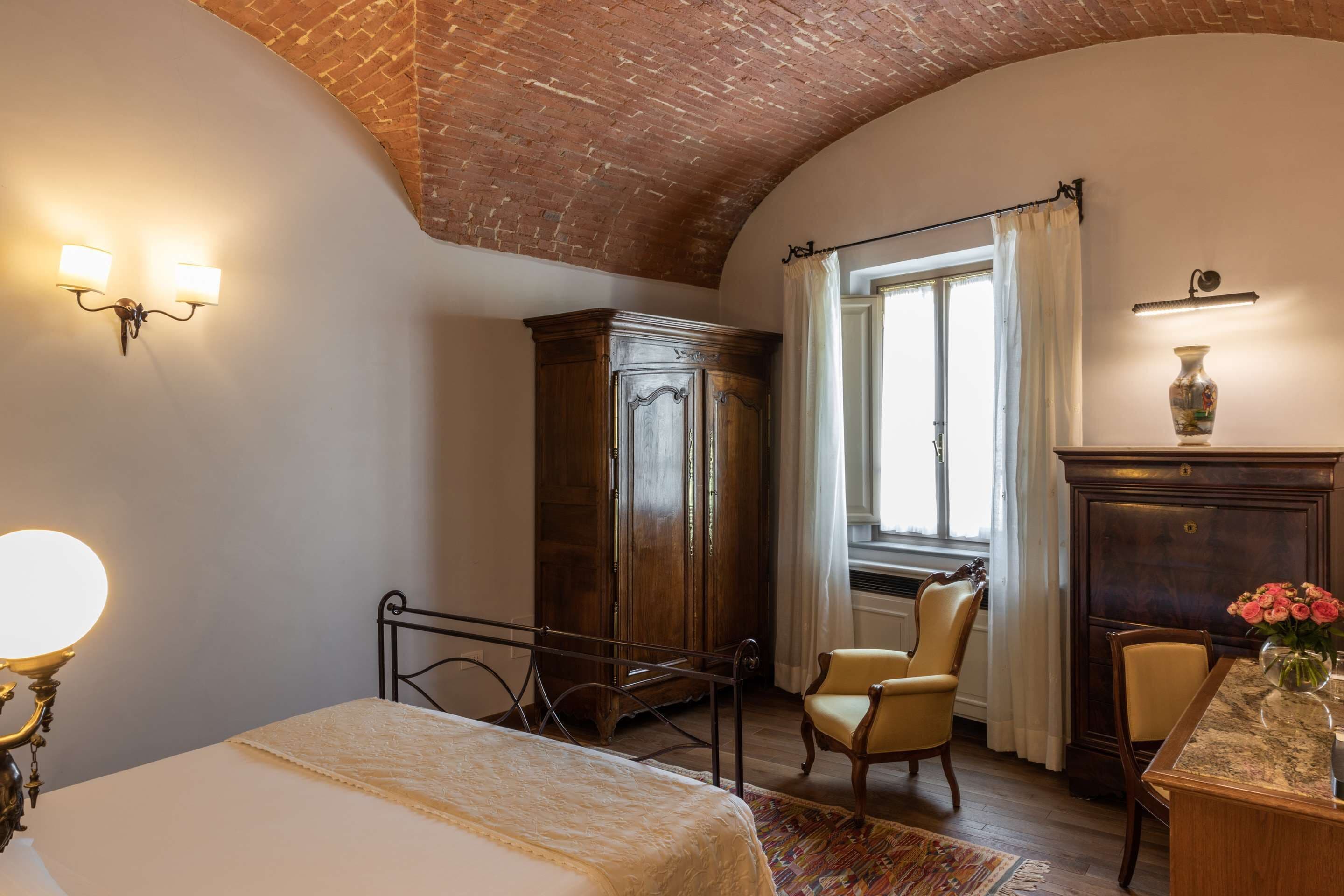 Un dormitorio con una cama, un armario de madera, una silla, un escritorio con un jarrón y una ventana con cortinas.