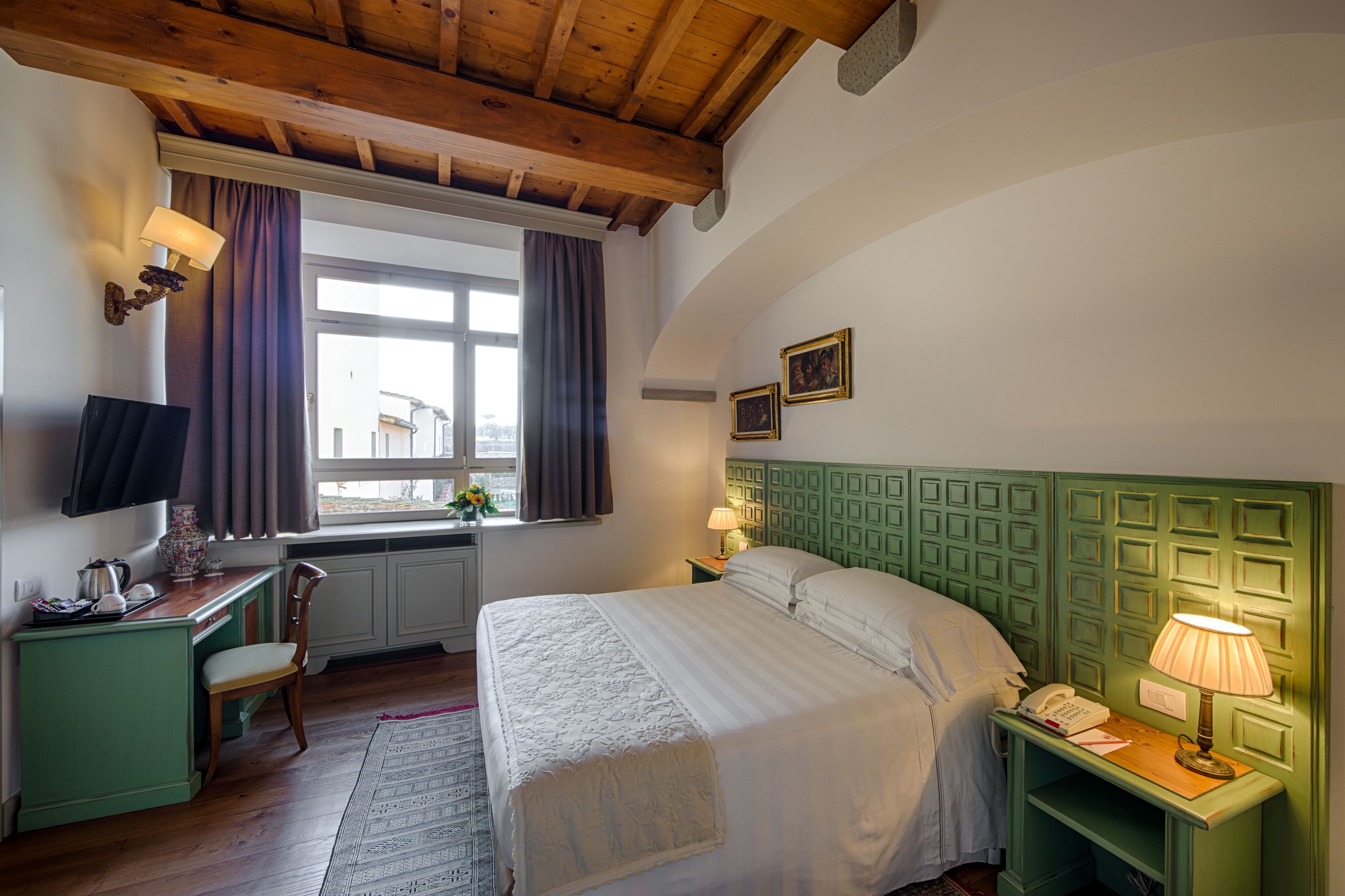 A hotel room with a bed, a desk with a lamp, a chair, a window with curtains, and a green headboard.