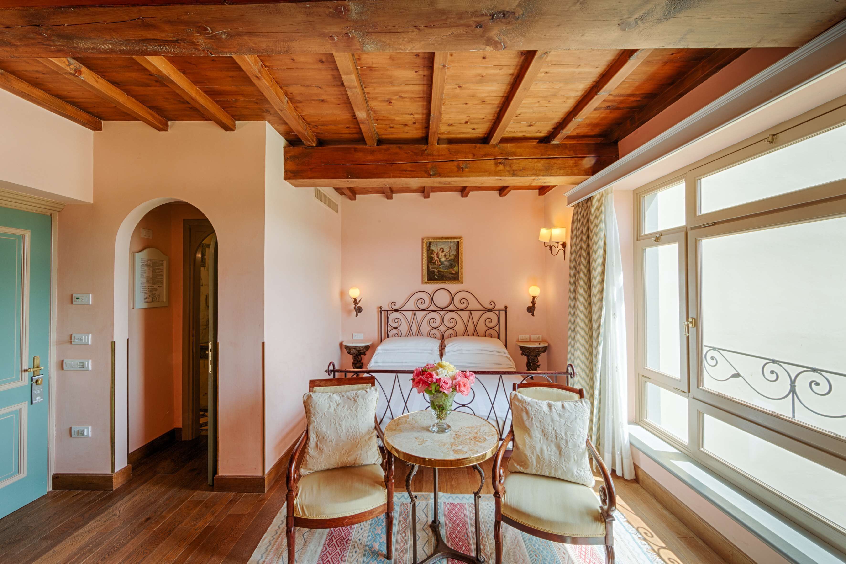 A bedroom with a wooden ceiling, a bed with a headboard, two bedside tables, a window with curtains, and a small table with chairs.