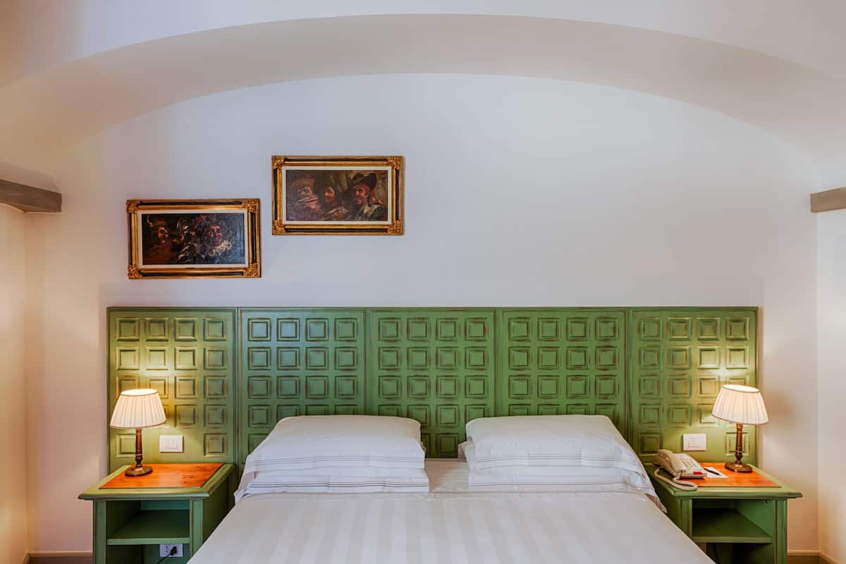 Habitación de hotel con cabecera verde, dos camas, lámparas de noche y dos cuadros en la pared.