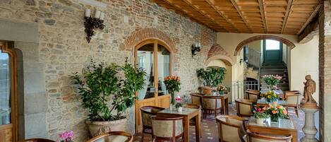 A stone-walled interior with wooden tables and chairs, potted plants, and a statue.