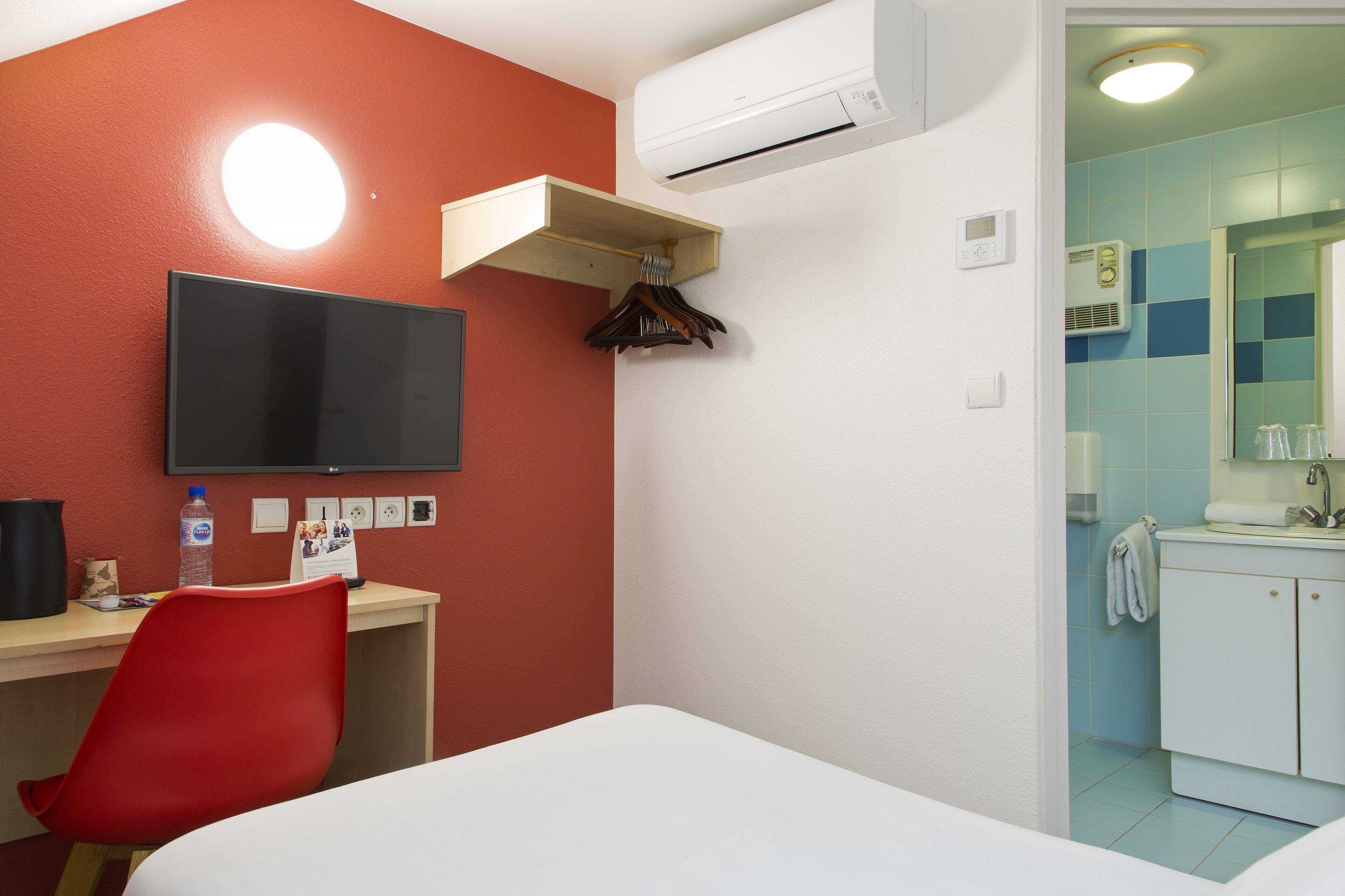 A hotel room with a bed, a desk with a chair, a television, and a bathroom with a sink and mirror.