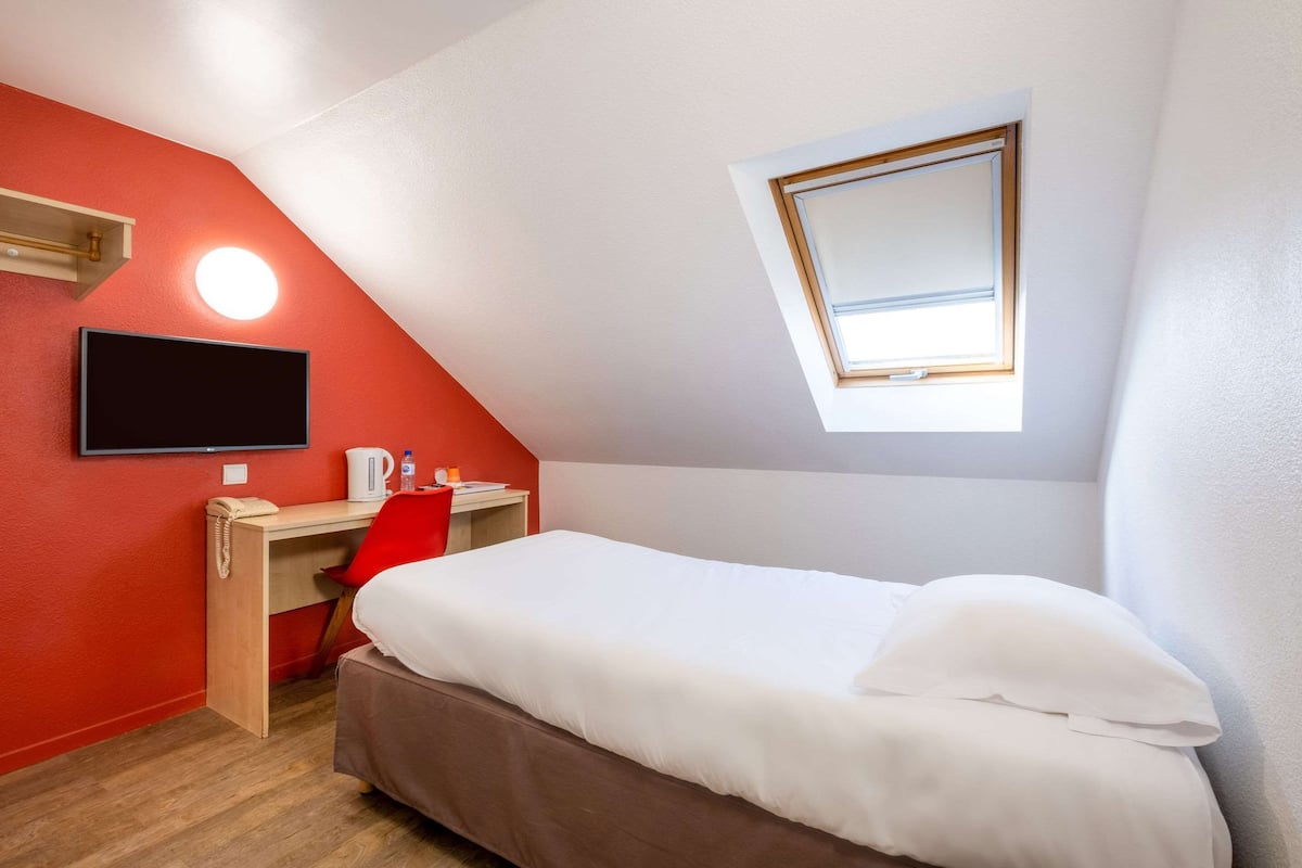 Une petite chambre moderne, équipée d’un lit, d’un bureau, d’une chaise, d’une télévision et d’une fenêtre.