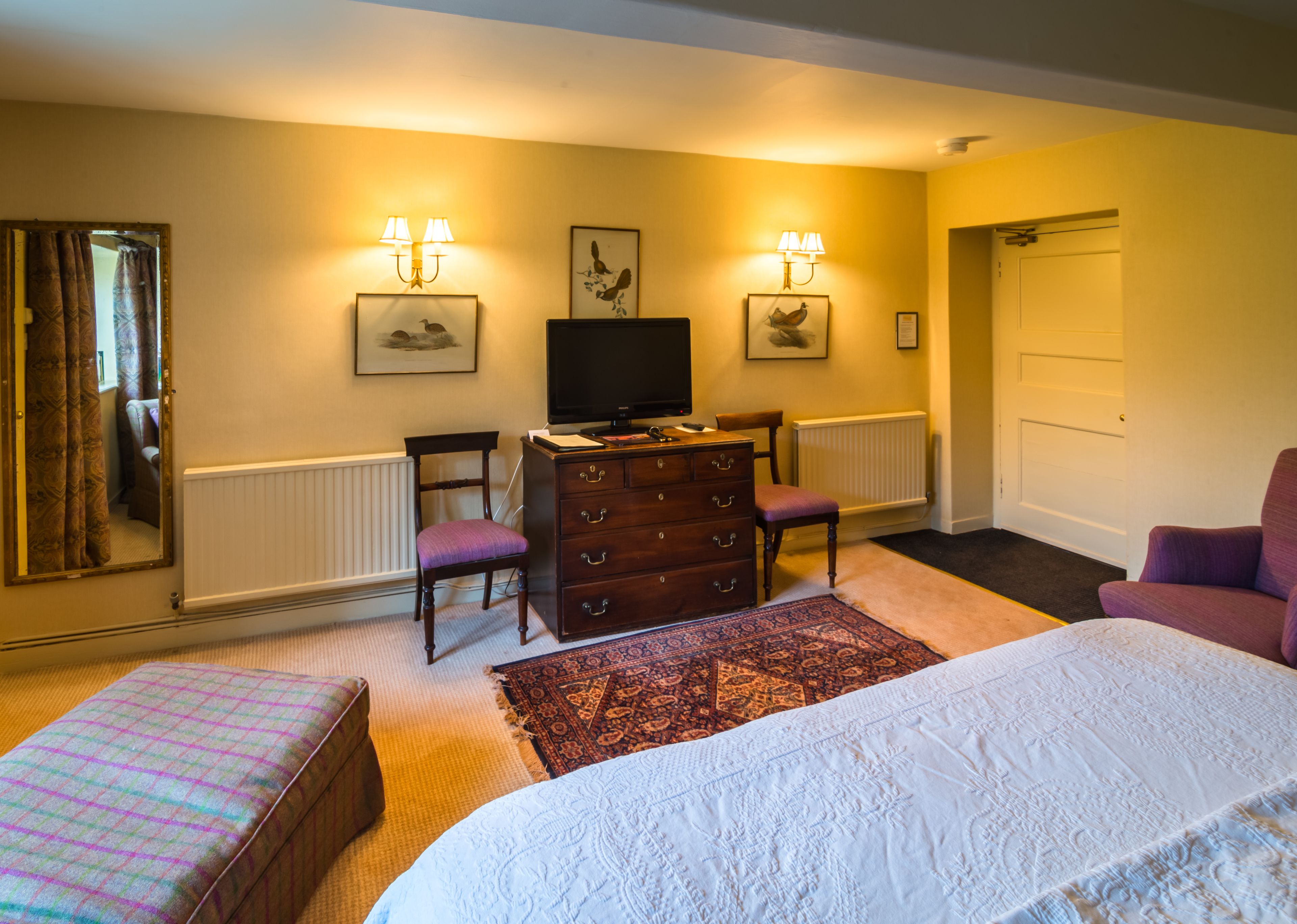 A hotel room with a bed, a dresser, a television, two chairs, a rug, a window with curtains, and a door.
