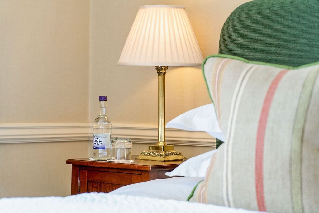 A neatly made bed with a green-striped pillow, a bedside table with a lamp, and a bottle of water.