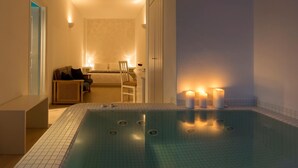 A modern indoor swimming pool with a tiled floor, a seating area with a chair and table, and candles on the poolside. - Tzekos Villas (Santorini)