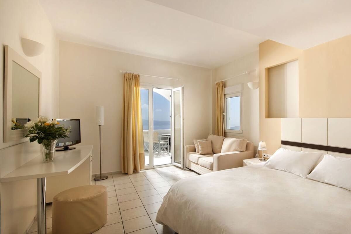 A hotel room with a large bed, a desk with a TV, a chair, and a balcony with curtains.