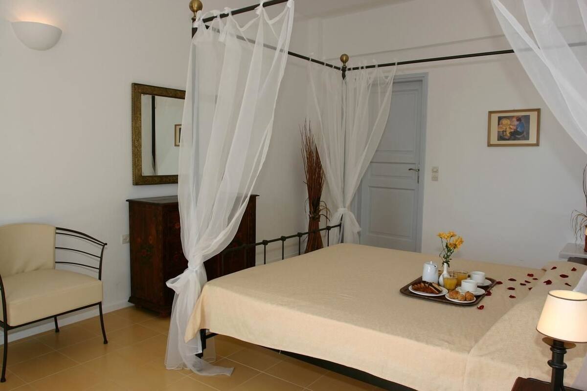 A bedroom with a four-poster bed, a bedside table with a lamp, a chair, a wooden cabinet, and a breakfast tray on the bed.