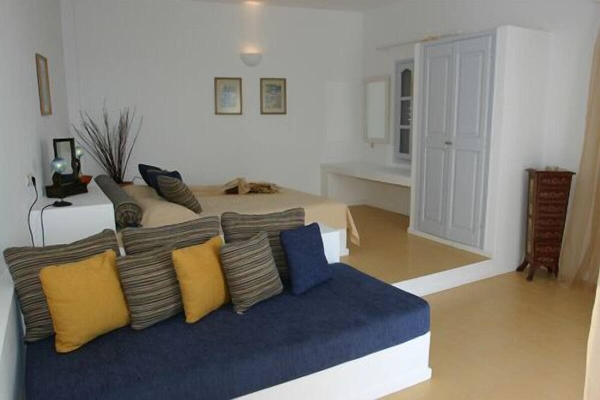 A modern living room with a sofa, a small table, and a bed. There are framed pictures on the wall and a door leading to another room.