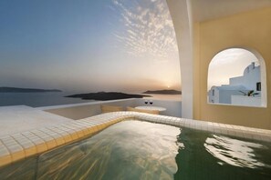 A serene indoor pool area with a view of a sunset over a body of water and a white building. - Tzekos Villas (Santorini)