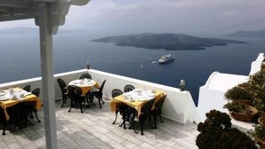 A terrace with tables set for dining, overlooking a body of water with a ship in the distance. - Tzekos Villas (Santorini)
