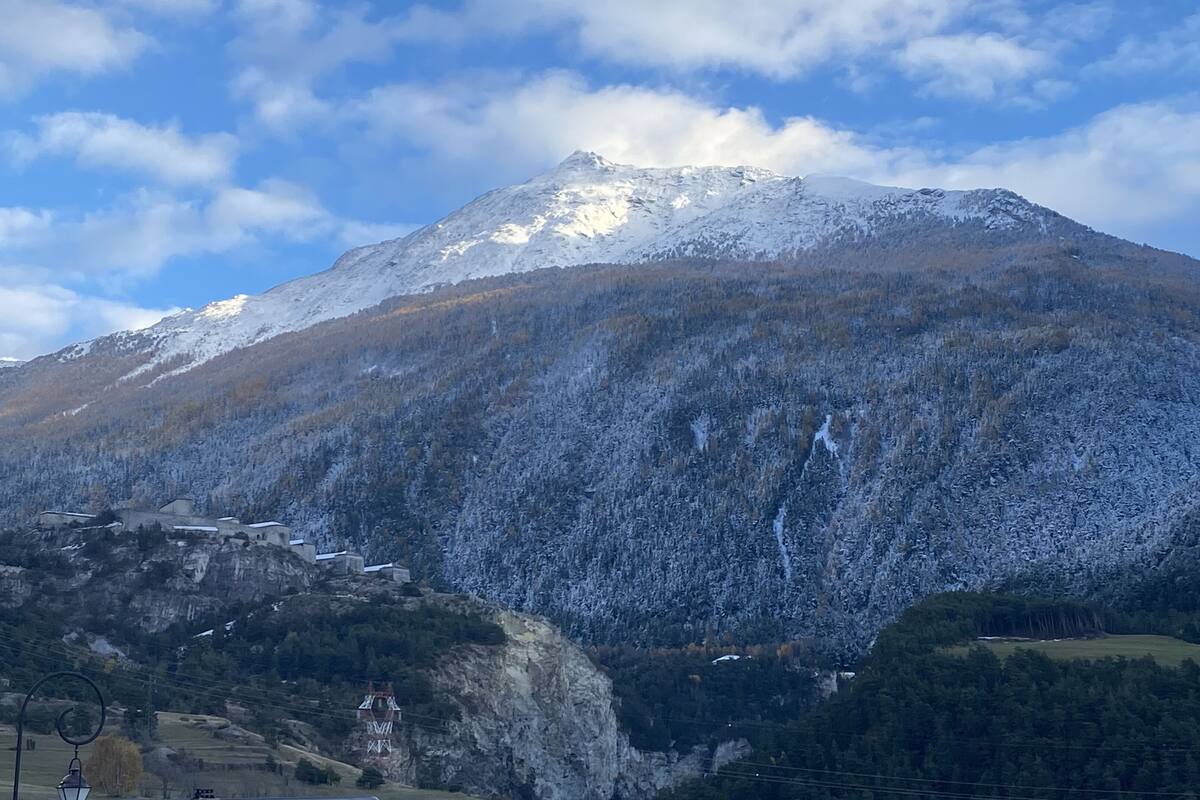 Un paysage montagneux avec des sommets enneigés, une vallée boisée et un petit village composé de bâtiments et d’une infrastructure de ligne électrique.