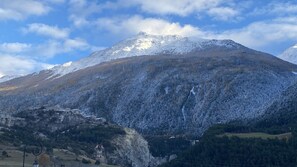 Paisaje montañoso con picos nevados, un valle boscoso y un pequeño asentamiento con edificios y una torre de líneas eléctricas.