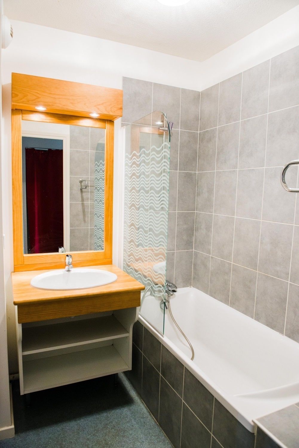 Un baño con tocador de madera, lavamanos blanco, espejo, ducha con puerta de cristal y paredes con azulejos grises.