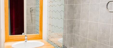 Un baño con tocador de madera, lavamanos blanco, espejo, ducha con puerta de cristal y paredes con azulejos grises.