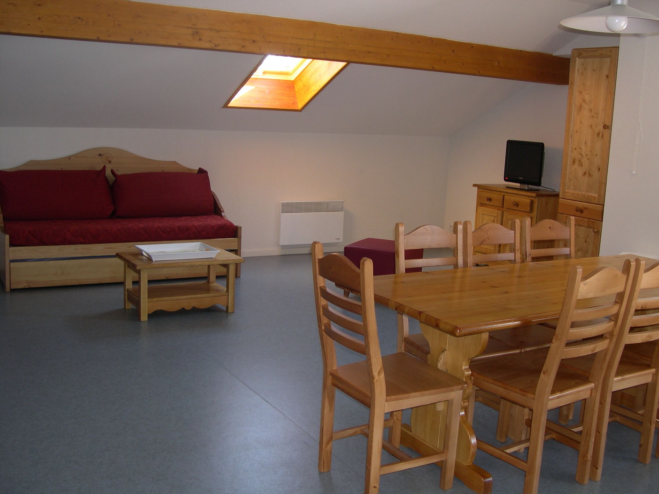 Een kamer met een houten eettafel, houten stoelen, een rode bank, een kleine houten tafel en een televisie op een houten kast.