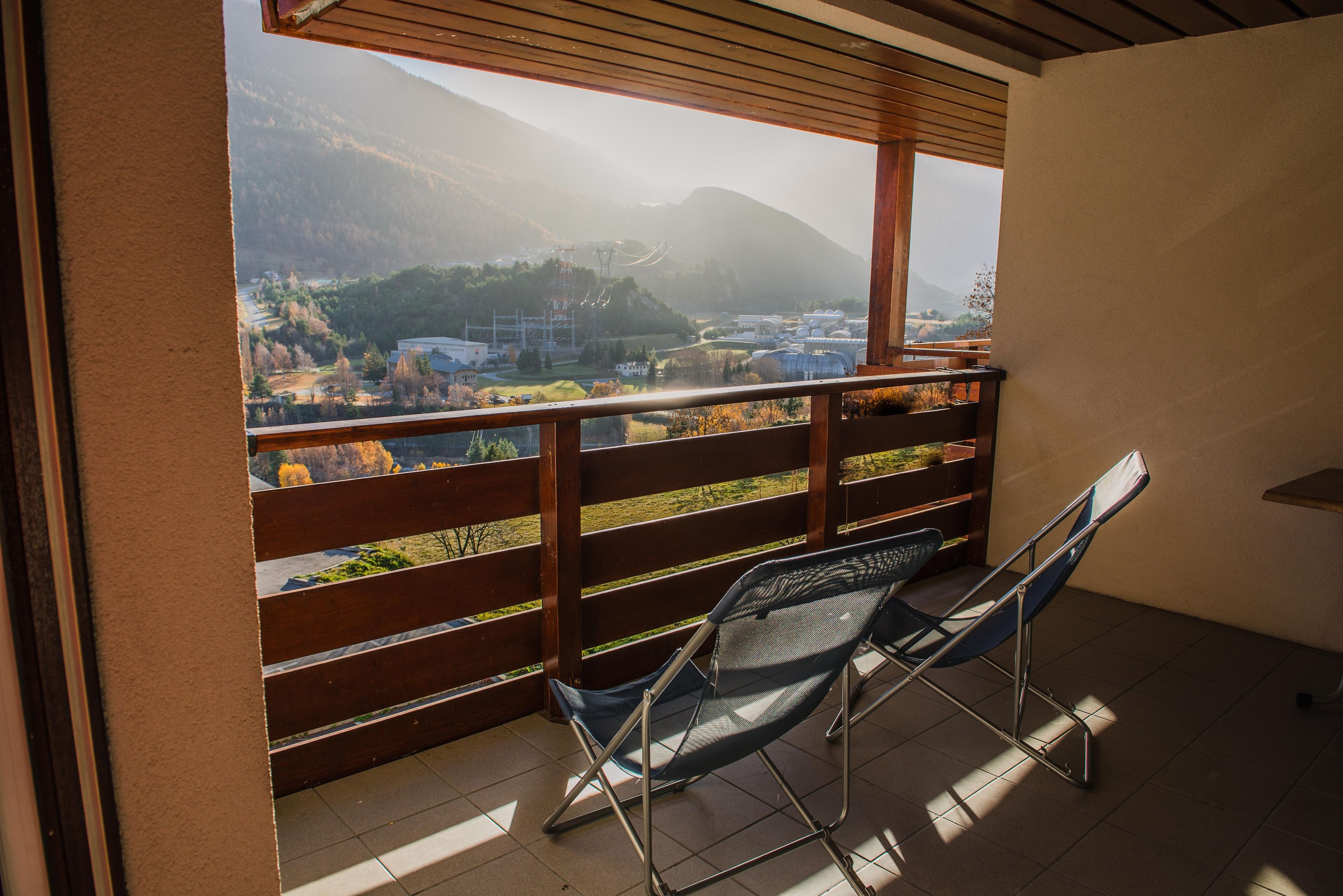 Un balcon doté d’une balustrade en bois, deux chaises pliantes bleues et une vue sur un paysage montagneux avec des bâtiments visibles au loin.