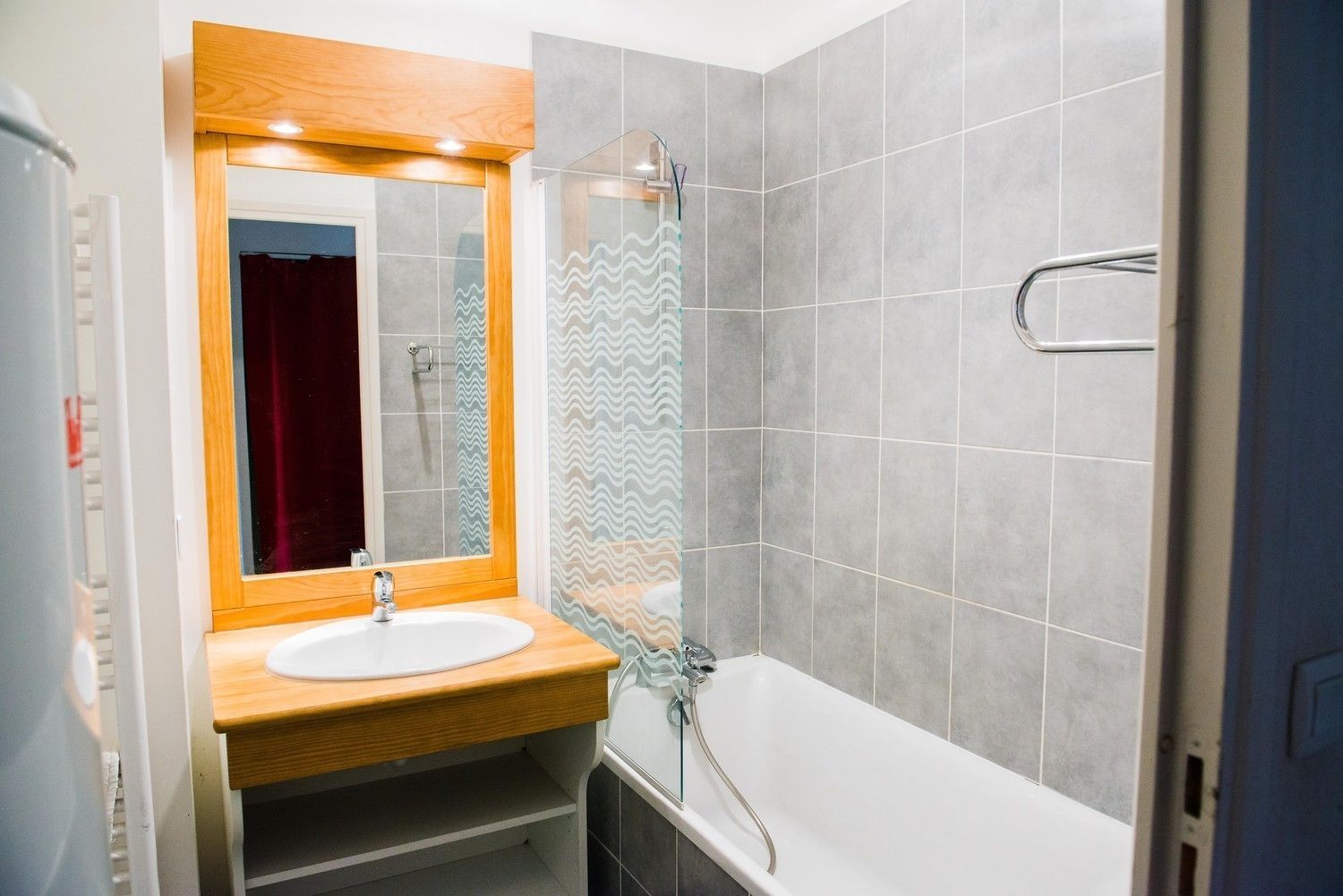 A bathroom with a wooden vanity, a white sink, a mirror, a shower with a glass door, and grey tiled walls.