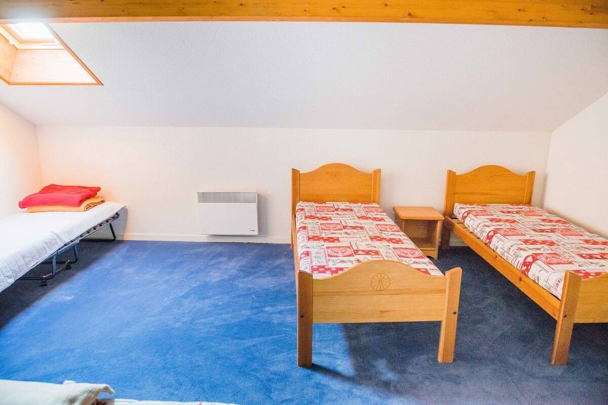 Un petit logement composé d’une seule pièce, comprenant deux lits en bois, une moquette bleue, un radiateur blanc et une lucarne.
